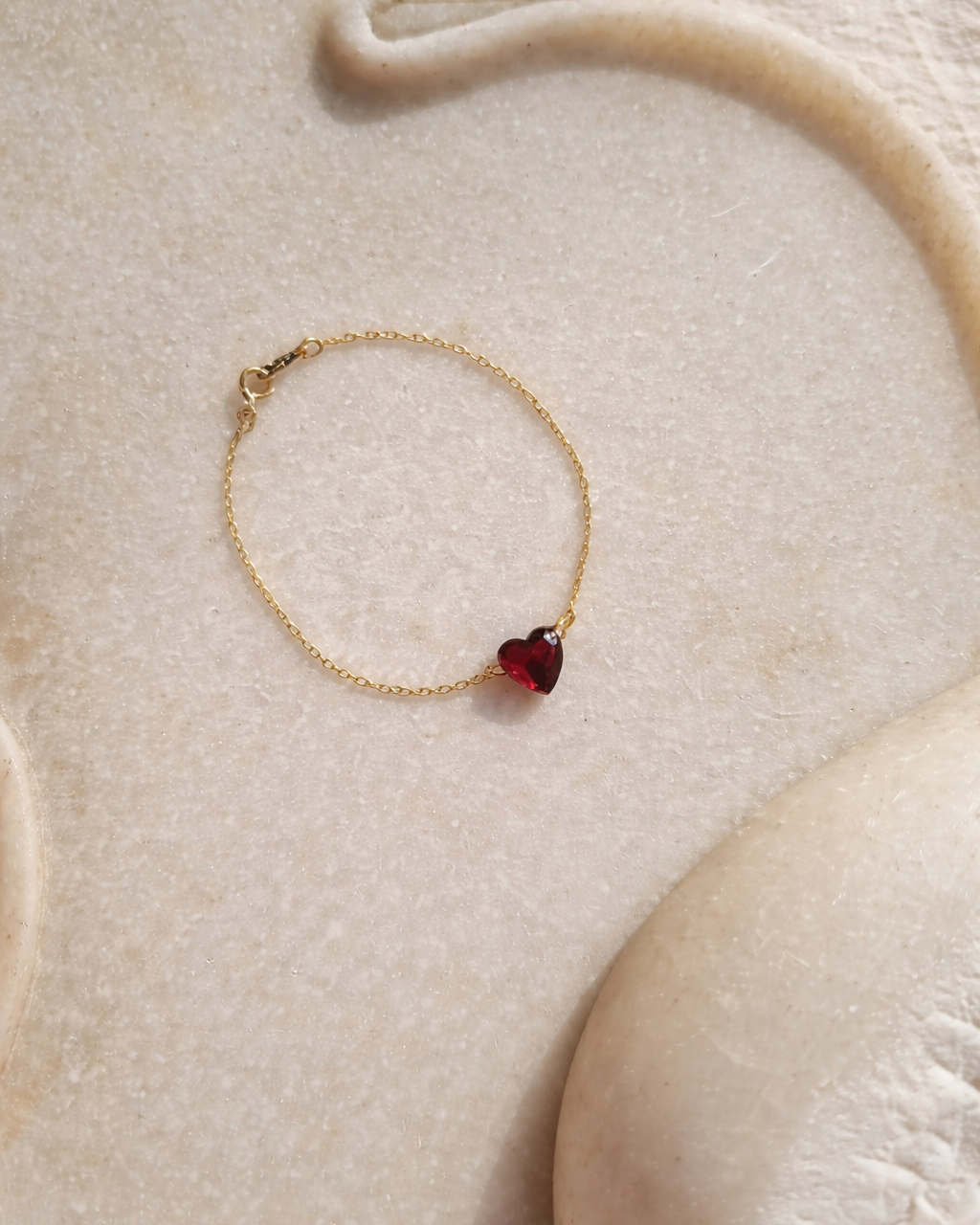 Elegant gold bracelet with red heart gemstone charm styled on neutral beige background, minimal luxury jewelry looks