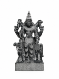 A dark gray stone statue of a multi-armed Hindu deity, likely Shiva, standing on a rectangular base against a plain white background.