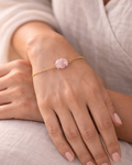 Hand wearing a gold bracelet with a pink gemstone on a light background