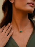 Woman wearing a gold necklace with a green pendant, touching her shoulder.