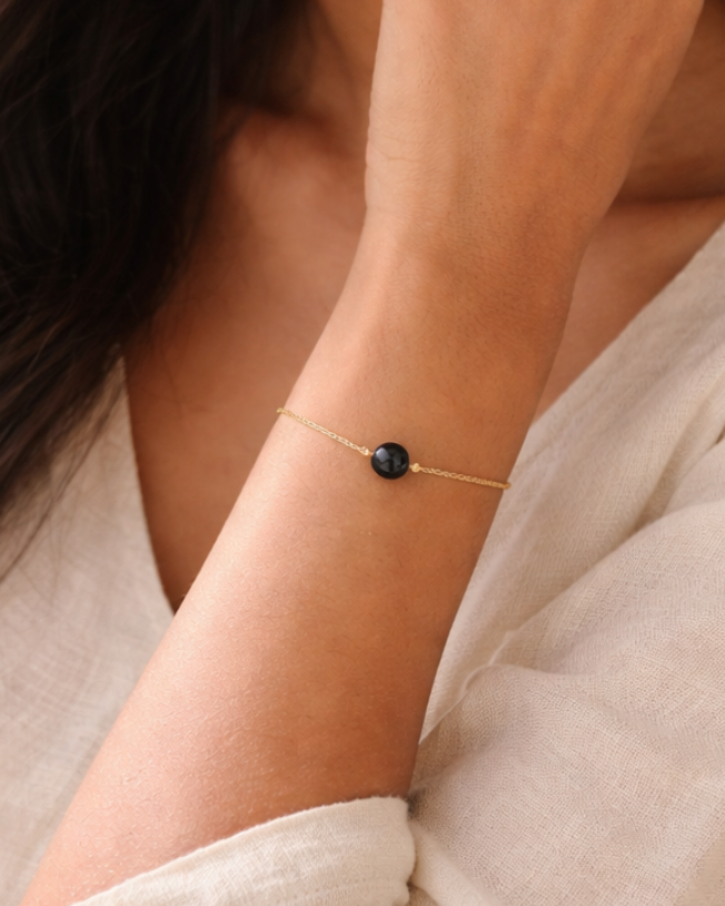 Woman wearing a delicate bracelet with a small black bead, against a neutral background.