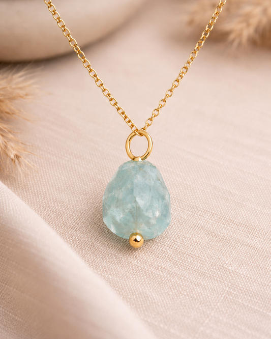 A gold chain necklace with a teardrop-shaped light blue faceted stone pendant and a small gold bead accent, displayed on a soft beige fabric background.