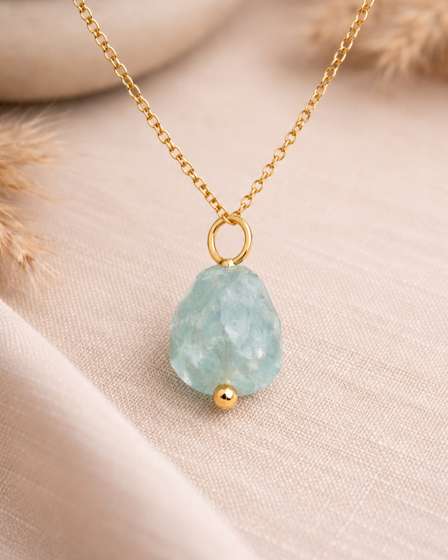A gold chain necklace with a teardrop-shaped light blue faceted stone pendant and a small gold bead accent, displayed on a soft beige fabric background.