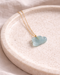 Aquamarine cloud pendant on a gold chain placed on a neutral beige ceramic plate, minimal aesthetic jewelry