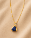 A gold chain necklace with a triangular pendant featuring a deep blue lapis lazuli stone, displayed on a soft beige fabric background.