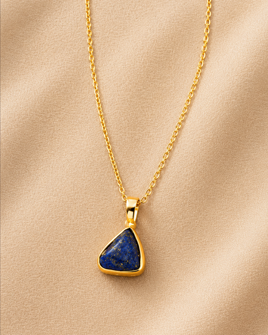 A gold chain necklace with a triangular pendant featuring a deep blue lapis lazuli stone, displayed on a soft beige fabric background.