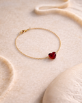 Minimal gold bracelet with red heart gemstone charm placed on a soft beige textured surface, elegant aesthetic jewelry