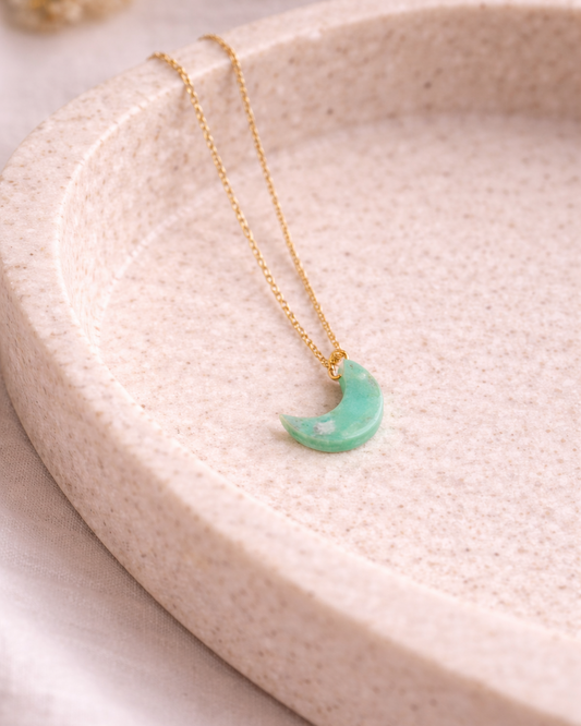 Malachite luna pendant on gold chain placed on a soft beige ceramic surface, minimal elegant gemstone jewelry