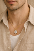 Man wearing a gold necklace with a circular pendant, dressed in a beige shirt.